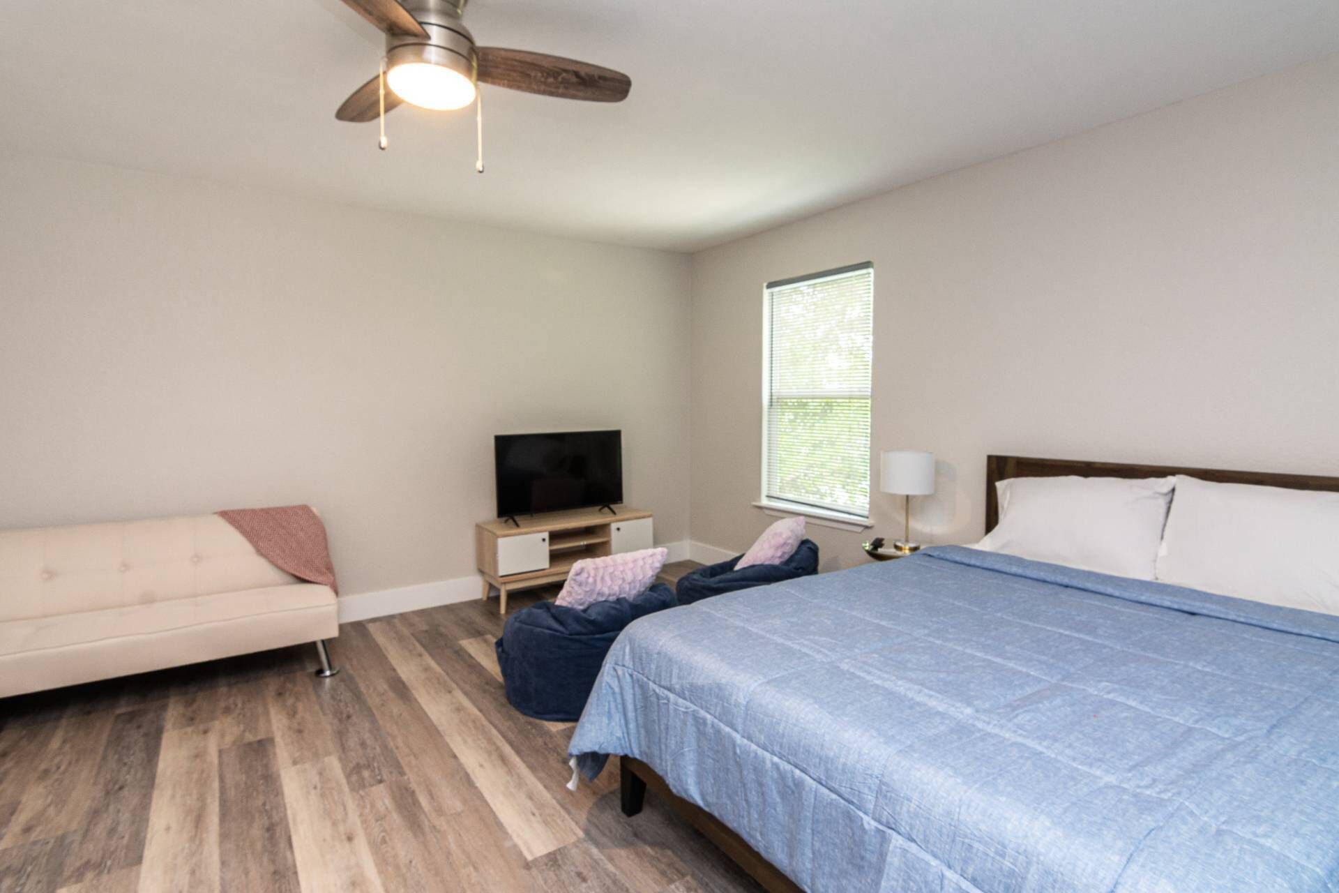 A bedroom with a bed, a TV on a wooden stand, a sofa, and a window with blinds.
