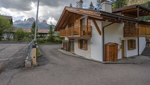 A two-story chalet with wooden balconies, a stone facade, and a view of mountains in the background. - Beautiful and modern apartment in the center of Alba di Canazei (Canazei)