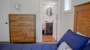 Quarto com penteadeira de madeira, cama com roupa de cama azul e corredor que dá para um banheiro.