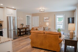 A modern kitchen with a stainless steel refrigerator, a dining area with a table and chairs, and a living room with a sofa and a lamp.