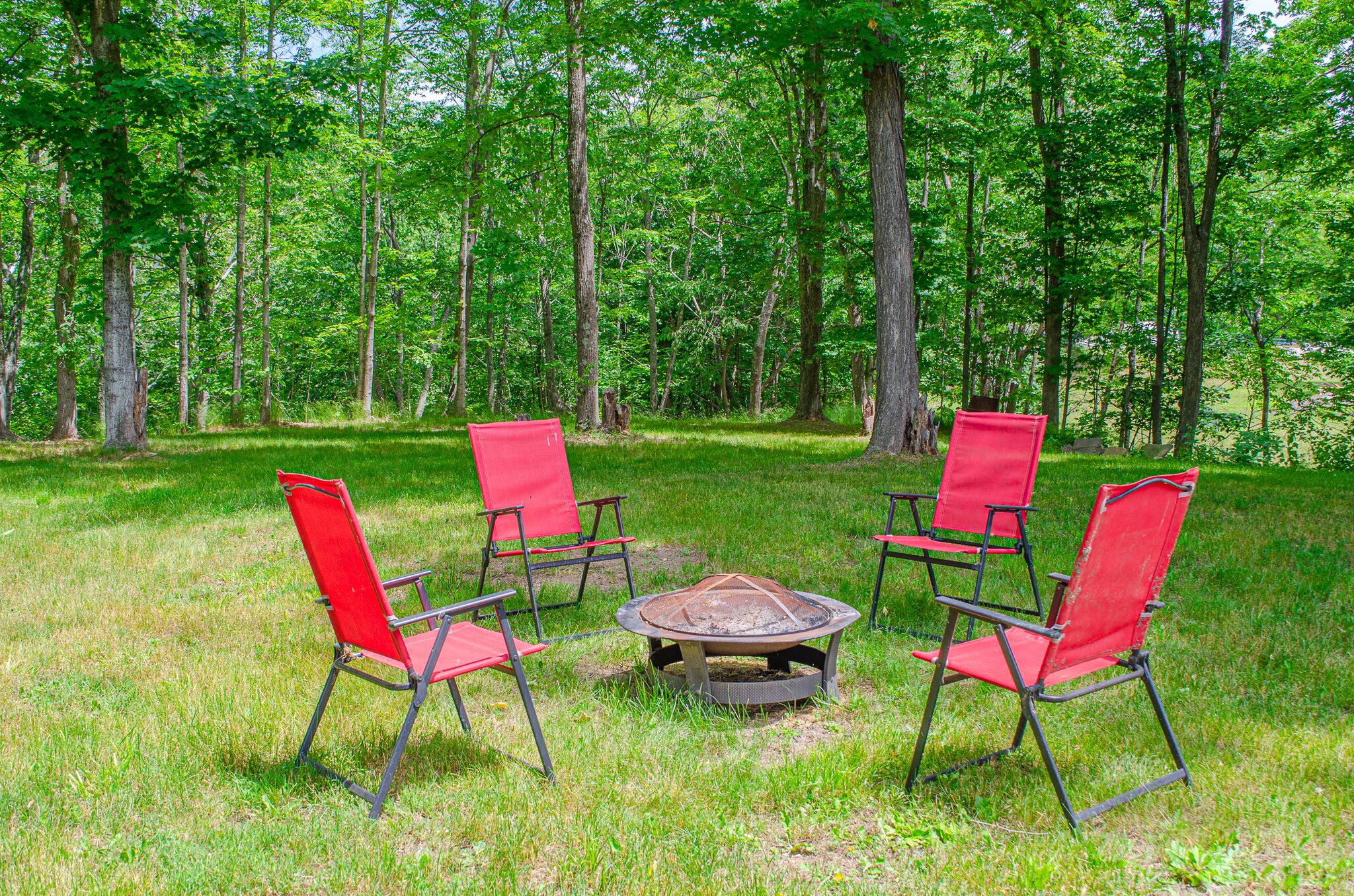 A circular fire pit surrounded by four red chairs in a grassy area with trees in the background.