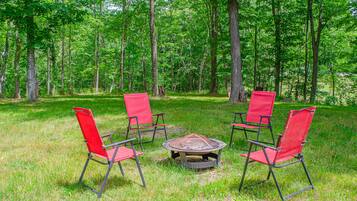 A circular fire pit surrounded by four red chairs in a grassy area with trees in the background.