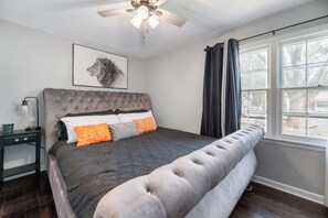Un dormitorio con cabecera capitonada, una cama grande con ropa de cama gris y naranja, una mesita de noche con lámpara y una ventana con cortinas.