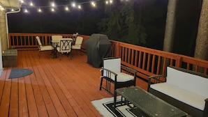 An outdoor deck with wooden flooring, white and black outdoor furniture, and string lights. - The Ultimate Sunset on Flathead Lake (Polson)
