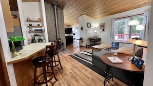 A cozy living room with a wooden ceiling, a dining area with wooden chairs, a kitchen with a stove, and a large window offering a view of the outdoors. - The Ultimate Sunset on Flathead Lake (Polson)