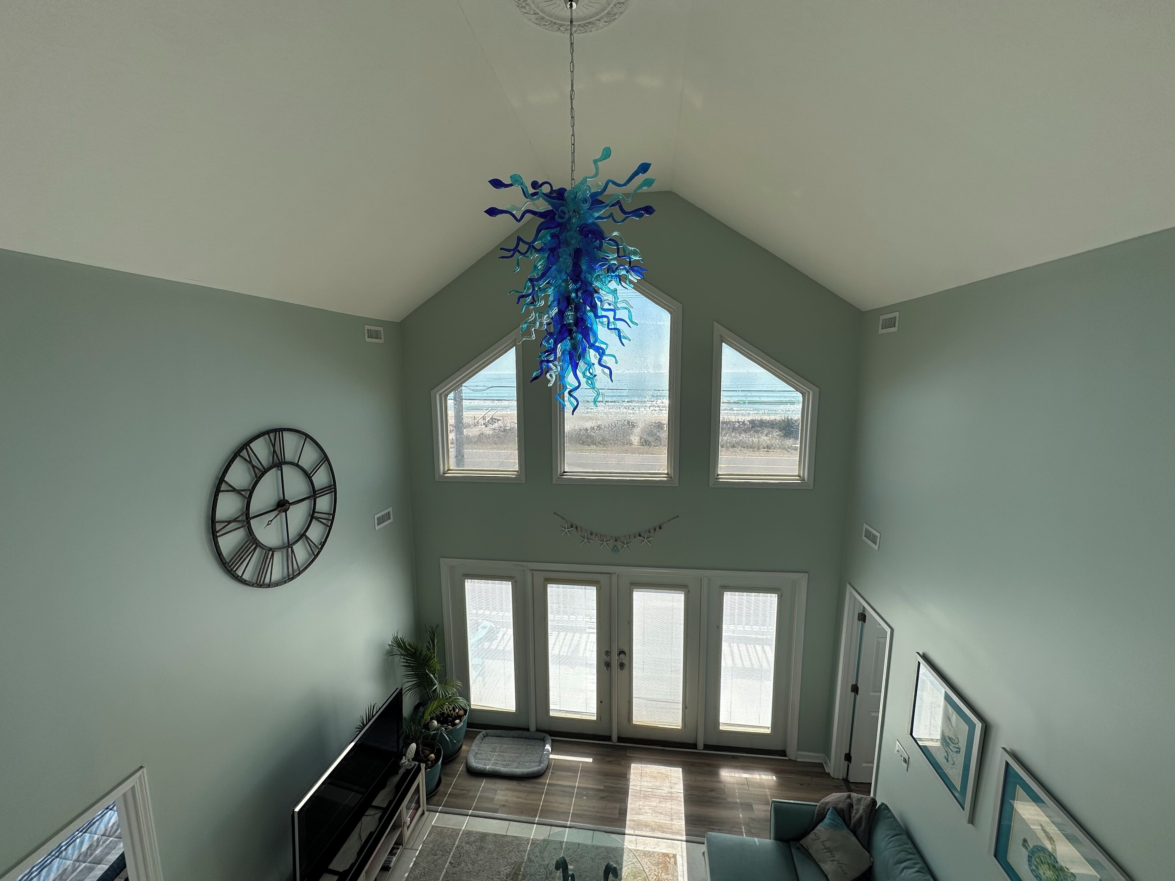 A spacious living room with a large window, a ceiling fan, and a decorative blue chandelier.
