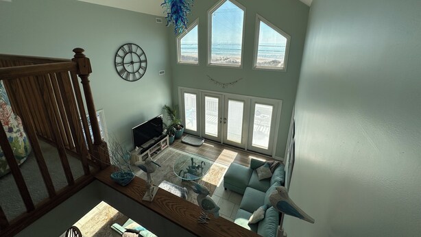 Un salón amplio con un ventanal grande que da a la playa, una escalera de madera y una lámpara moderna.