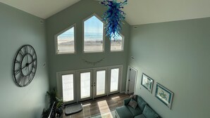 Amplia sala de estar con techo alto, ventanas grandes, una lámpara moderna y vistas al exterior.