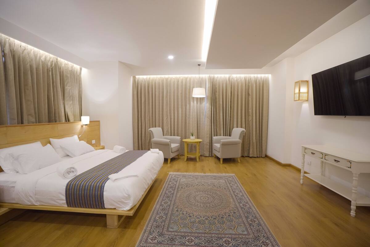 A modern hotel room with a large bed, two armchairs, a small table, a TV, and a patterned rug.