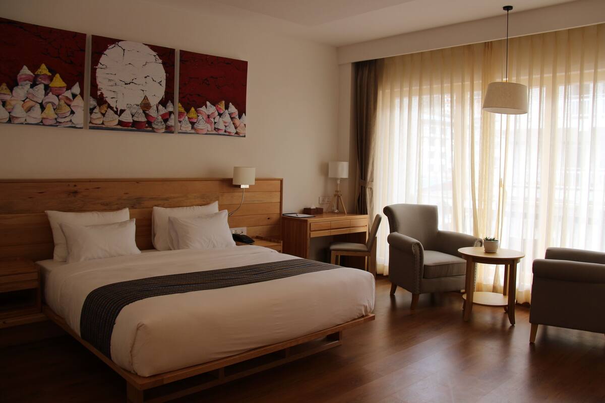 A hotel room with a large bed, a desk, two chairs, a small table, and a large window with curtains.