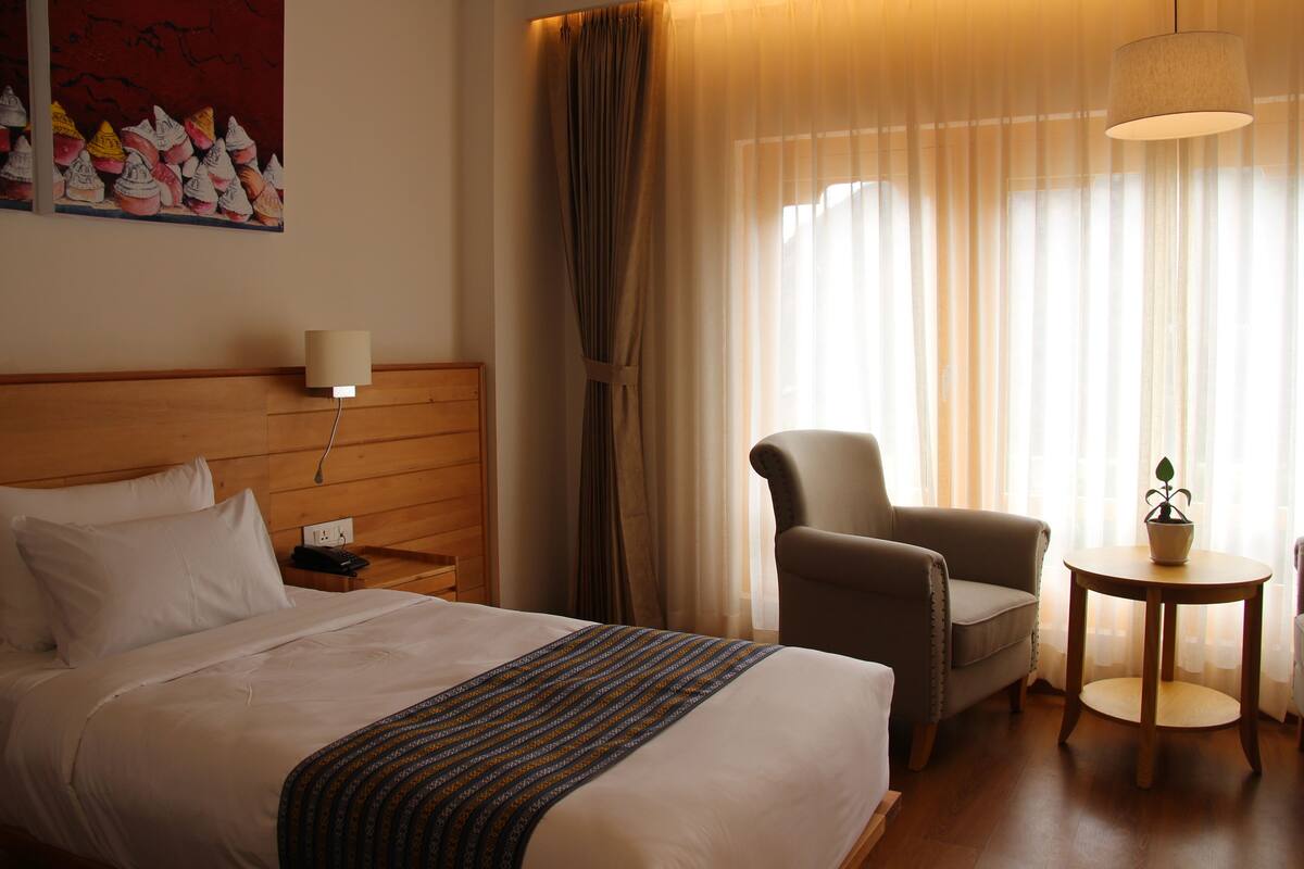A hotel room with a bed, a chair, a small table with a plant, and a painting on the wall.