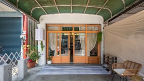 Entrada de un edificio con un porche cubierto, puerta corredera de vidrio y un pequeño rincón de descanso con una silla y una mesa.