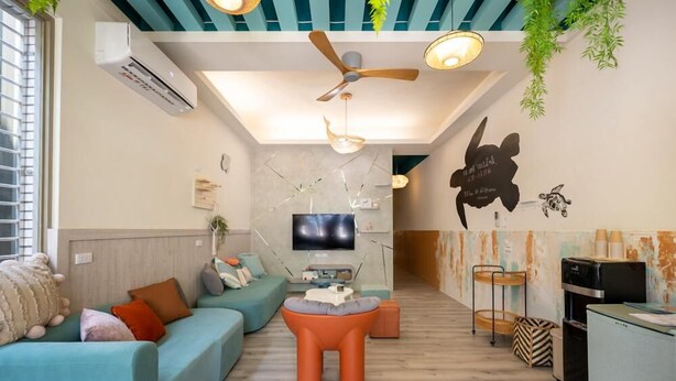 Una sala de estar moderna con sofá, reposapiés, ventilador de techo, televisor y una zona de cocina con refrigerador y estufa.