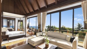 A spacious living area with a large sofa, a coffee table, and a bed. There are large windows offering a view of the ocean.