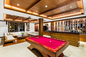 A pool table in a well-lit room with wooden ceiling beams, a bar area with bottles, and a seating area with a sofa and a coffee table.