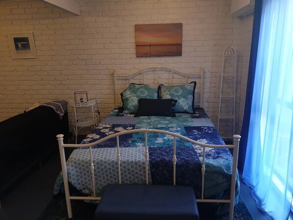 Une chambre avec un mur en briques blanches, un lit recouvert d’une courtepointe à motifs floraux bleus et blancs, un oreiller noir et un tableau représentant un coucher de soleil.