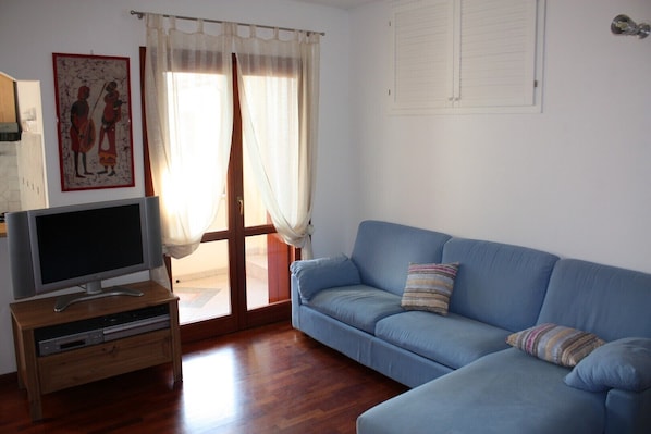Ein Wohnzimmer mit einem blauen Sofa, einem Flachbildfernseher auf einem Holzboden und einer Tür mit transparenten Vorhängen.