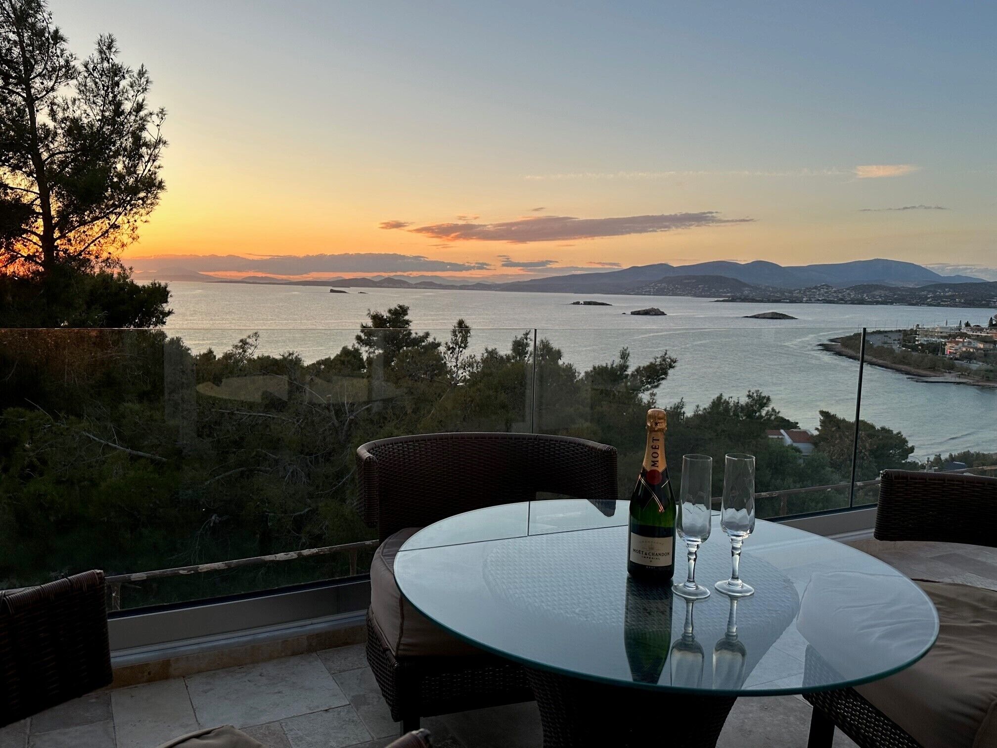 A glass table with a bottle of Moet & Chandon champagne and two glasses, overlooking a scenic view of a body of water and distant mountains.