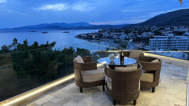 A balcony with wicker furniture and a glass table, overlooking a coastal town and sea. - Luxurious 2-bed Seaside Apartment in Saronida (Saronikos)