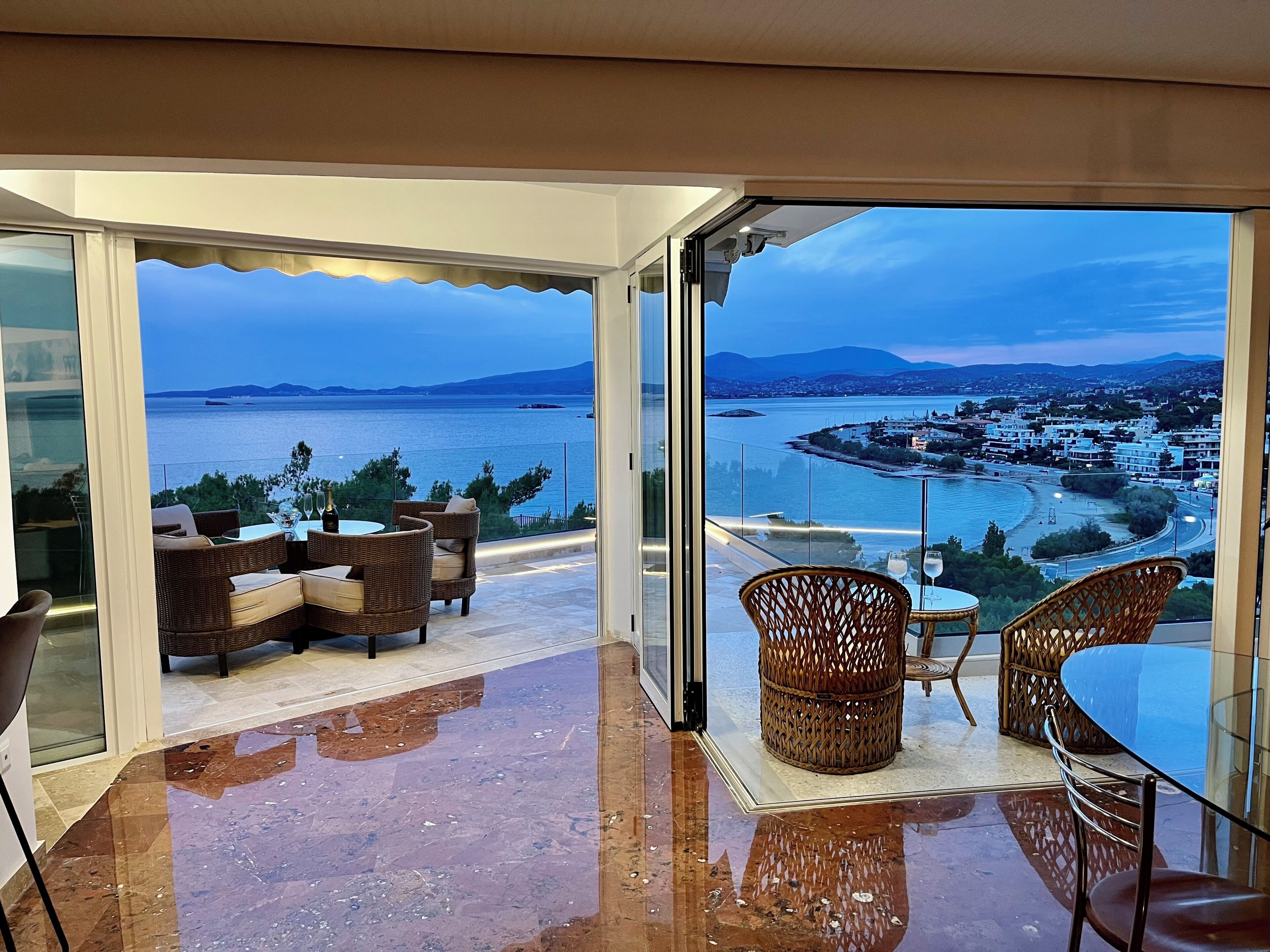 A balcony with wicker furniture, a glass table, and a view of the sea and town.