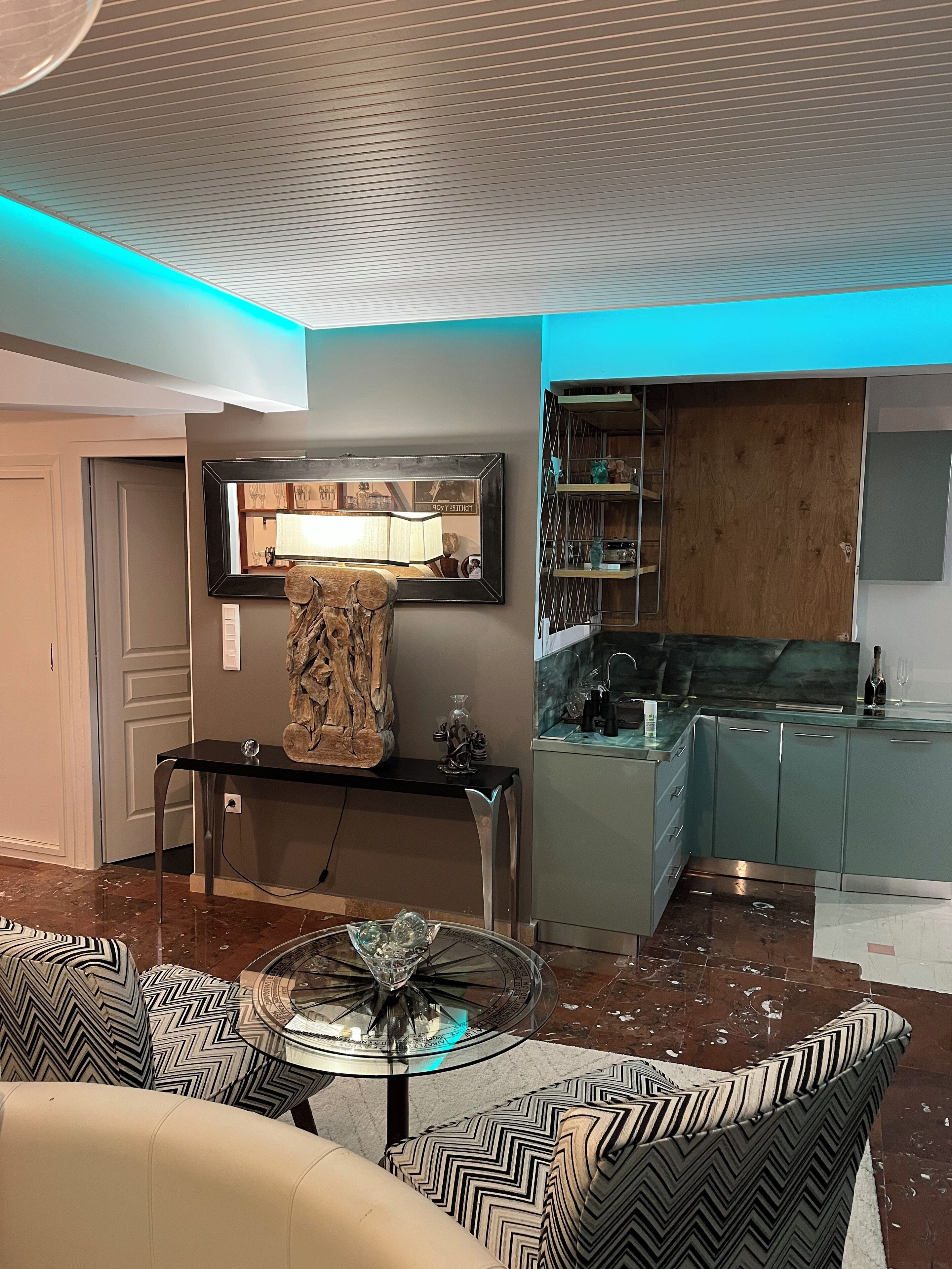 A modern living room with a glass coffee table, patterned sofas, a wooden sculpture, and a kitchen area with a wooden cabinet and a wine bottle.