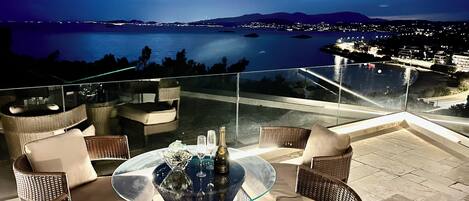 A glass-top table with wicker chairs and a bottle of champagne, overlooking a body of water and city lights at night.