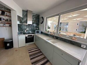 A modern kitchen with a built-in oven, microwave, and sink. There is a window with a view of buildings outside. - Luxurious 2-bed Seaside Apartment in Saronida (Saronikos)