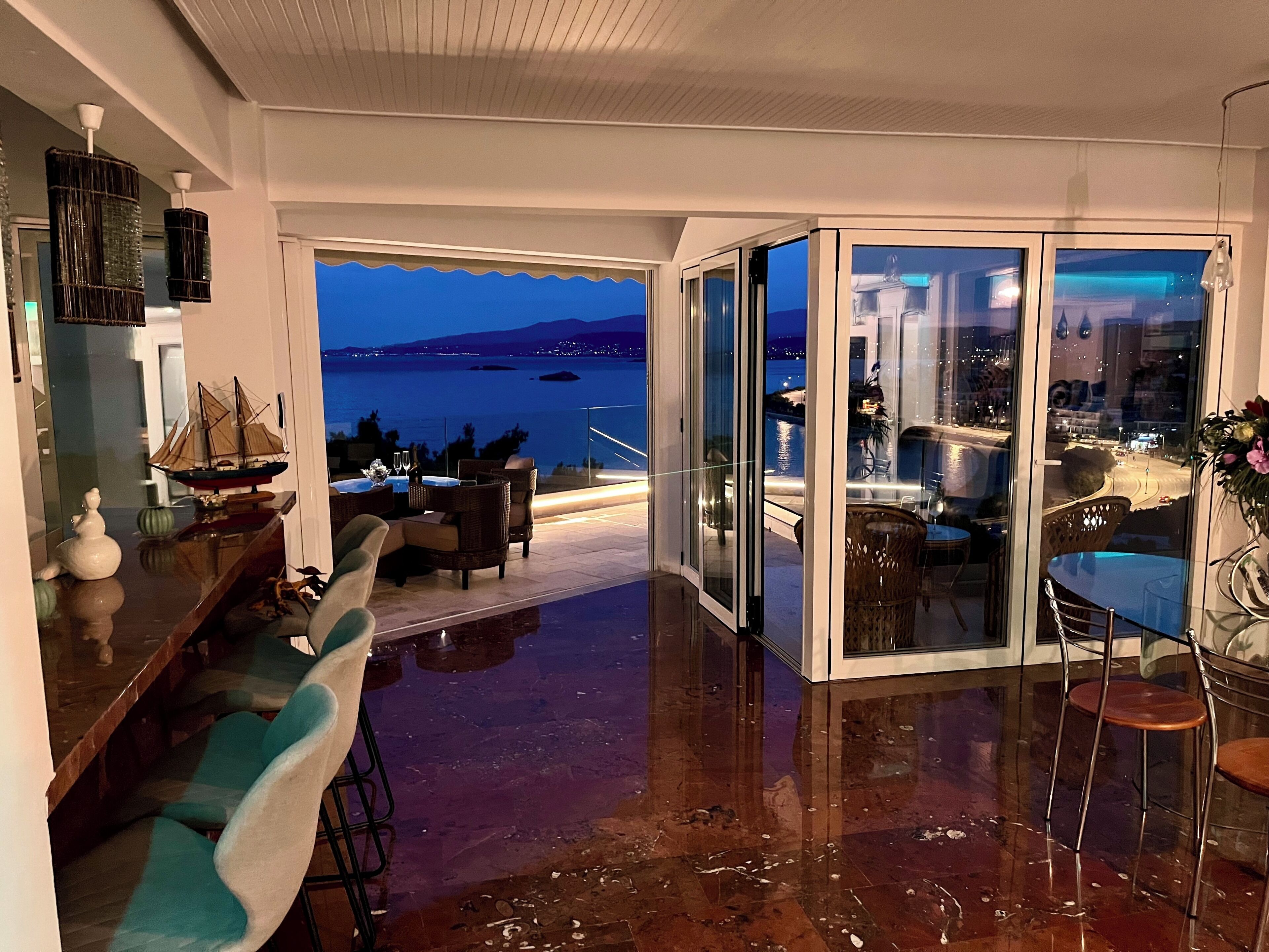 A modern living room with a view of the sea, a dining area with a table and chairs, and a glass-enclosed balcony.