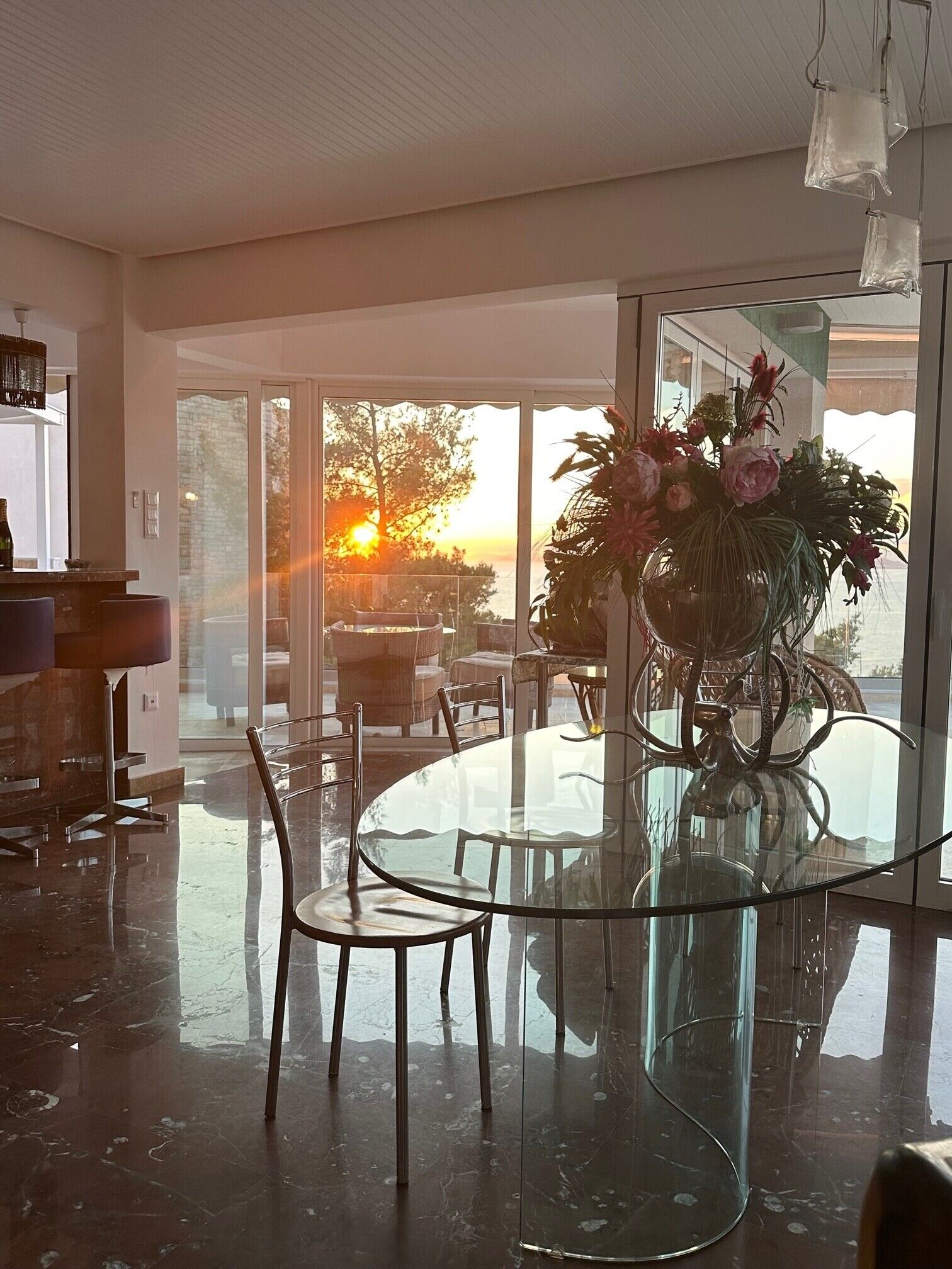 A modern dining area with a glass table and chairs, a vase of flowers, and a view of the sunset through large windows.