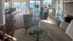 A spacious living room with a large sofa, a glass coffee table, and a dog. There are multiple seating areas and a view of the sea through large windows. - Luxurious 2-bed Seaside Apartment in Saronida (Saronikos)