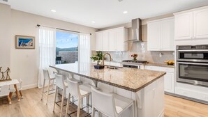 A modern kitchen with white cabinetry, a granite island with bar stools, stainless steel appliances, and a large window with a view. - Resort-like residence with pool, jacuzzi, sauna, ocean breeze & amazing sunsets (Mission Viejo)