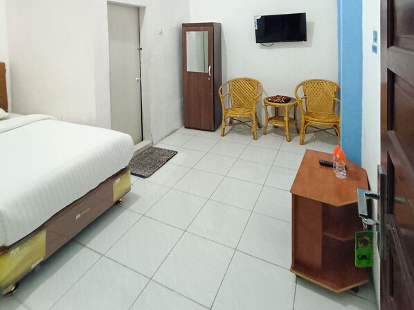 A hotel room with a bed, a desk, two wicker chairs, a small table with snacks, a television, and a door.