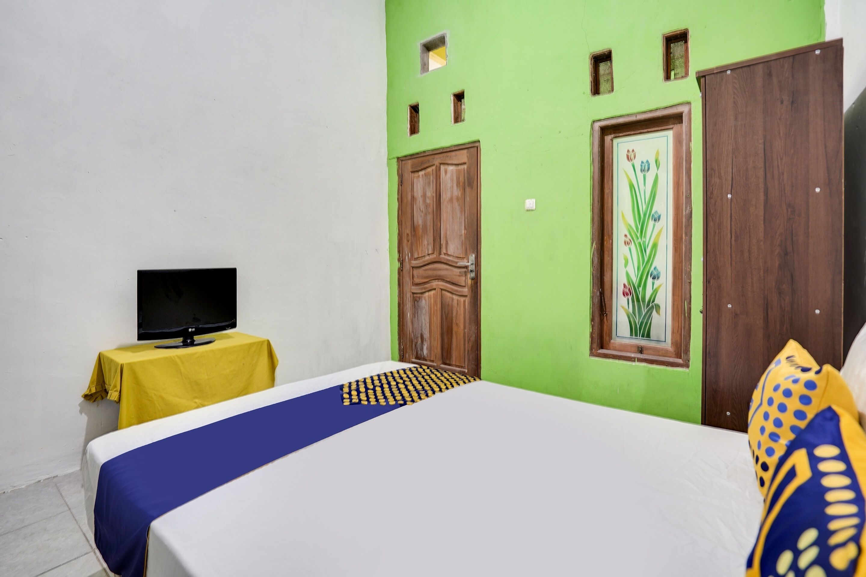Una habitación con una cama, un televisor sobre una mesita y una pared verde con una puerta y una ventana.