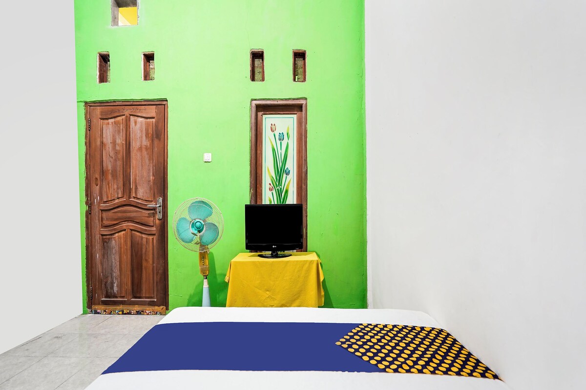 A room with a green wall, a wooden door, a fan, a TV on a yellow table, and a bed with a blue and yellow patterned bedspread.