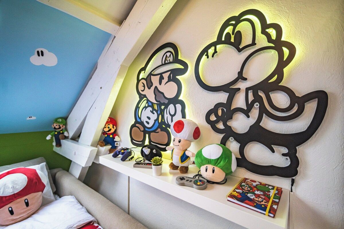 Une pièce avec une étagère blanche où sont exposés des peluches de Mario et Luigi, un coussin à l’effigie de Mario et un élément de décoration murale sur le thème de Mario.