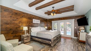 Une chambre avec un plancher en bois, un lit recouvert de draps blancs, un ventilateur de plafond, une télévision et une vue sur l’extérieur.