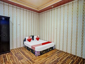 A bedroom with a wooden floor, a bed with patterned bedding, and a dark-colored wardrobe. - Hotel O Taman Wisata Iman Ii Near Taman Wisata Iman Sitinjo (Kota Sidikalang)