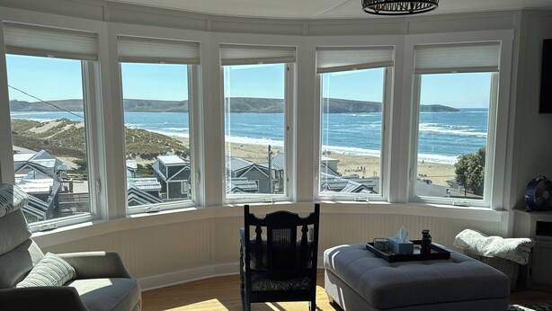 A room with large bay windows overlooking a beach, furnished with a sofa, ottoman, and a chair. - Unobstructed ocean views in a cozy setting (Dillon Beach)