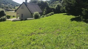 A small, single-story house with a sloped roof, surrounded by lush green fields and hills.