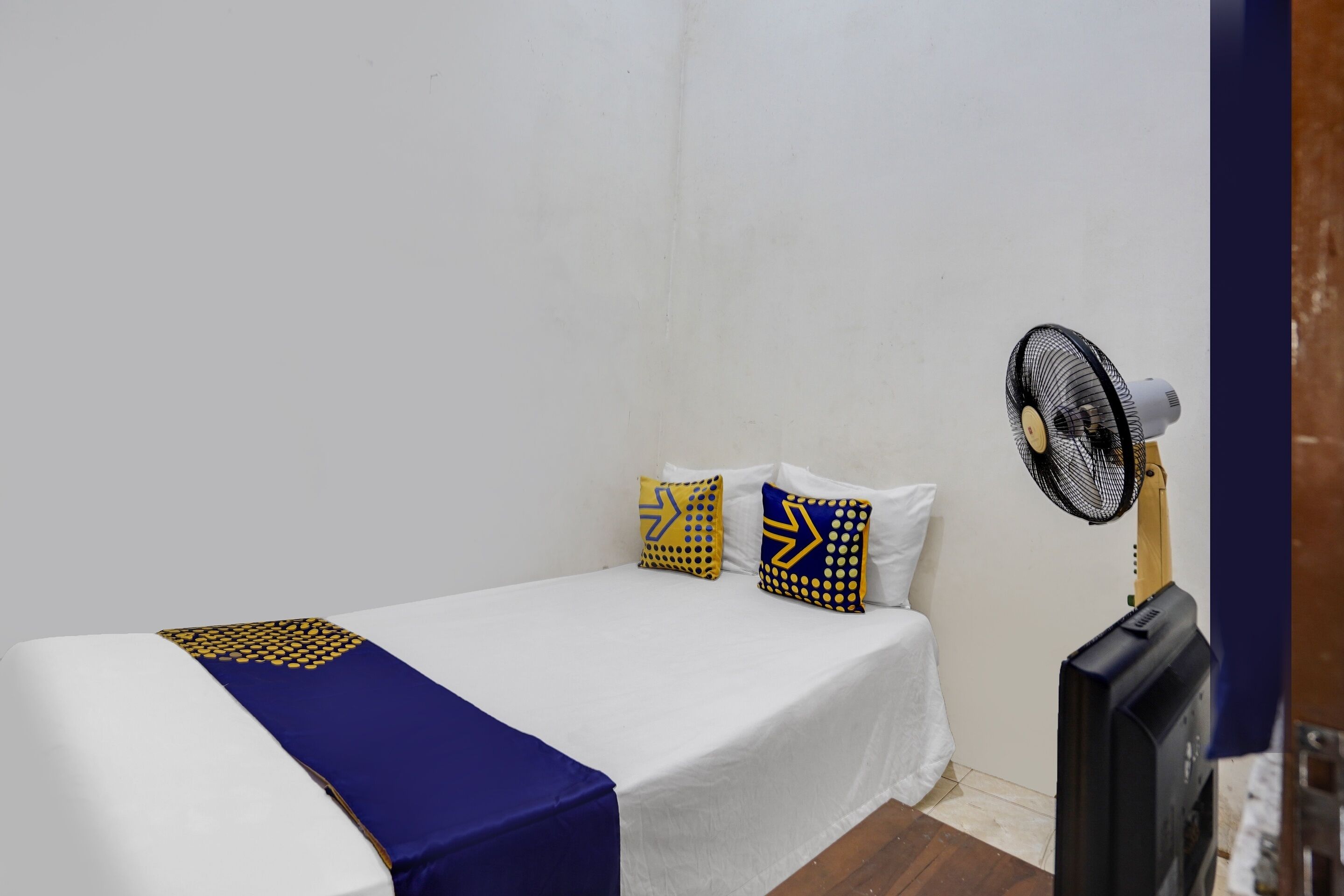 A small, single-bed room with a white bedspread, blue and yellow pillows, and a standing fan.