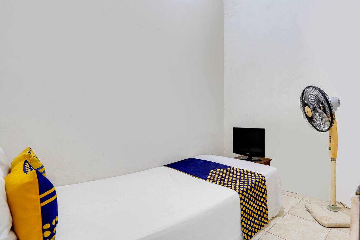 A small, single-bed room with a white bedspread, a yellow and blue pillow, a black television on a small table, and a standing fan.
