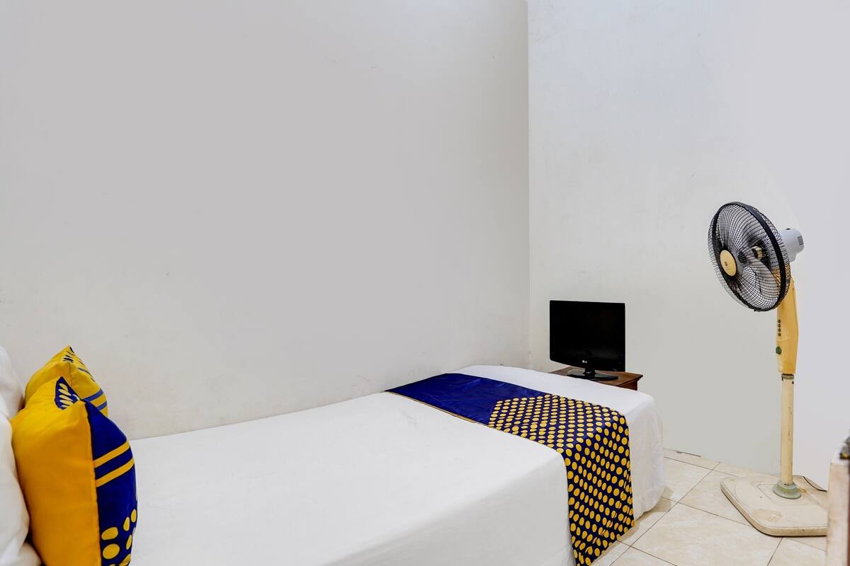 A single bed with a white mattress, a blue and yellow patterned blanket, and a yellow pillow with a blue design. A small table with a black object on it is next to the bed. A standing fan is positioned to the right of the bed.