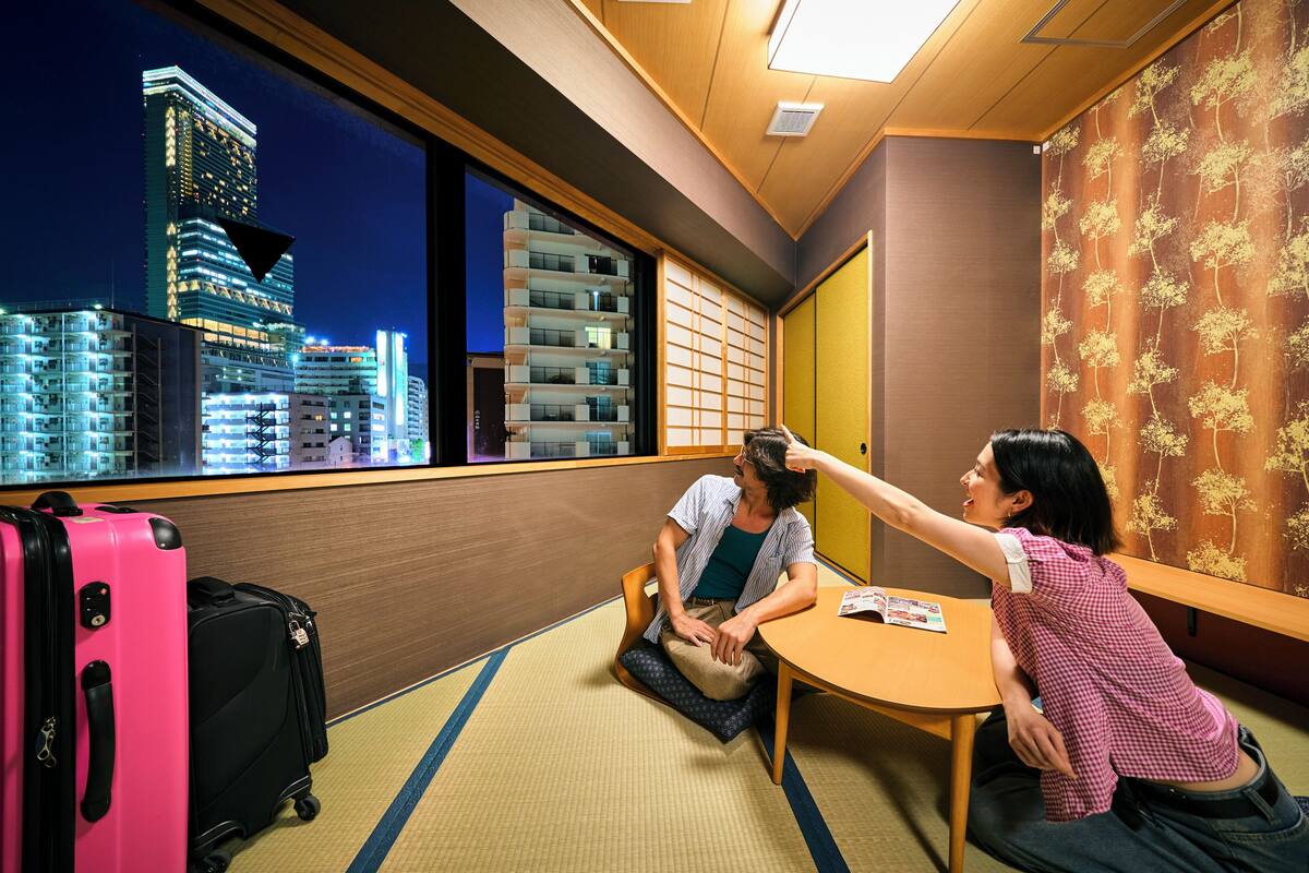 A hotel room with tatami flooring, a traditional Japanese sliding door, and a view of city buildings at night.