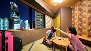 Une chambre d’hôtel avec un sol recouvert de tatamis, une porte coulissante de style japonais traditionnel et une vue sur les bâtiments de la ville la nuit.