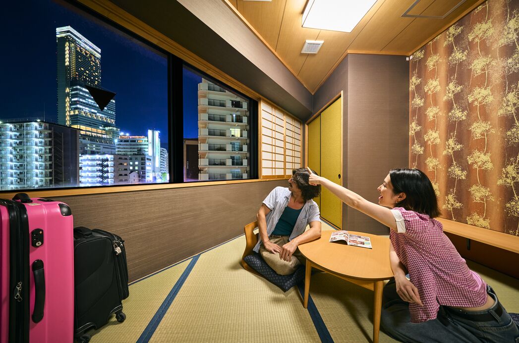 A hotel room with a city view, tatami mat flooring, a small table, and a bed.
