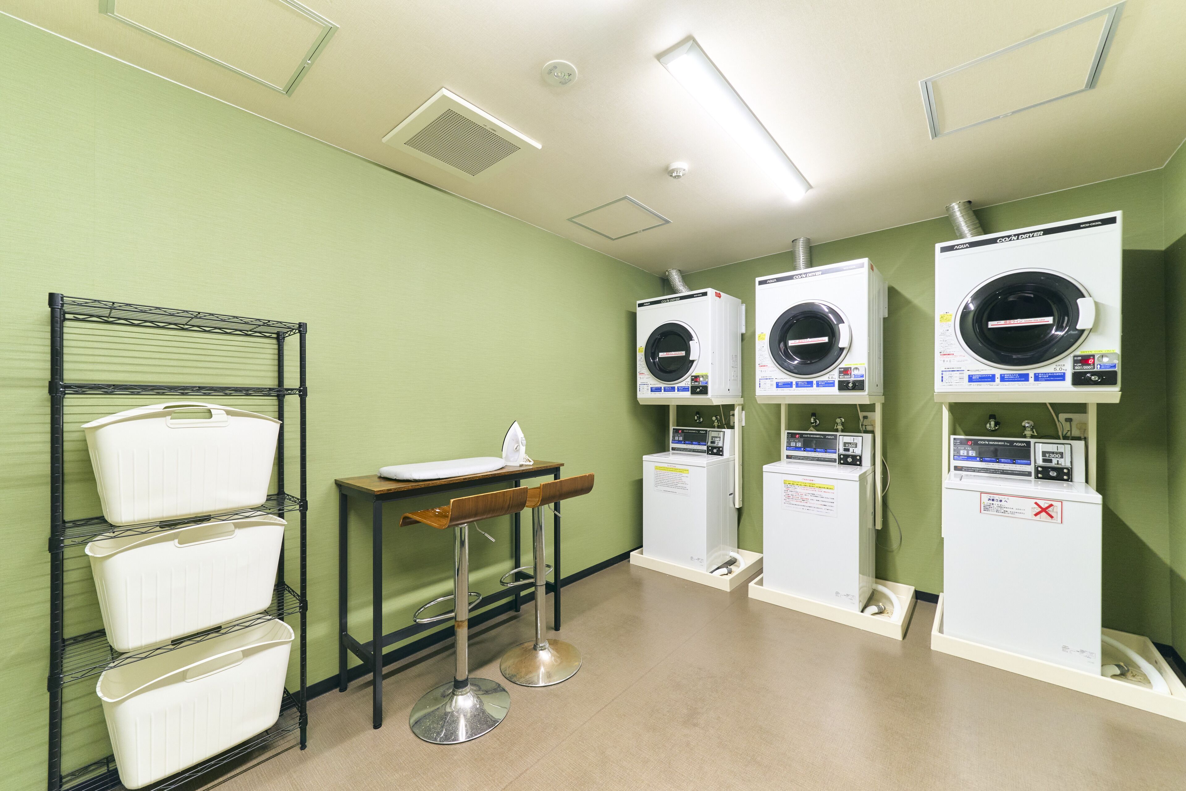 A laundry facility with multiple washing machines, a drying rack, and a small table with an ironing board.