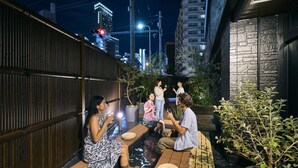 Un groupe de personnes est assis autour d’une table en bois à l’extérieur, la nuit. On aperçoit des bâtiments et une rue en arrière-plan.