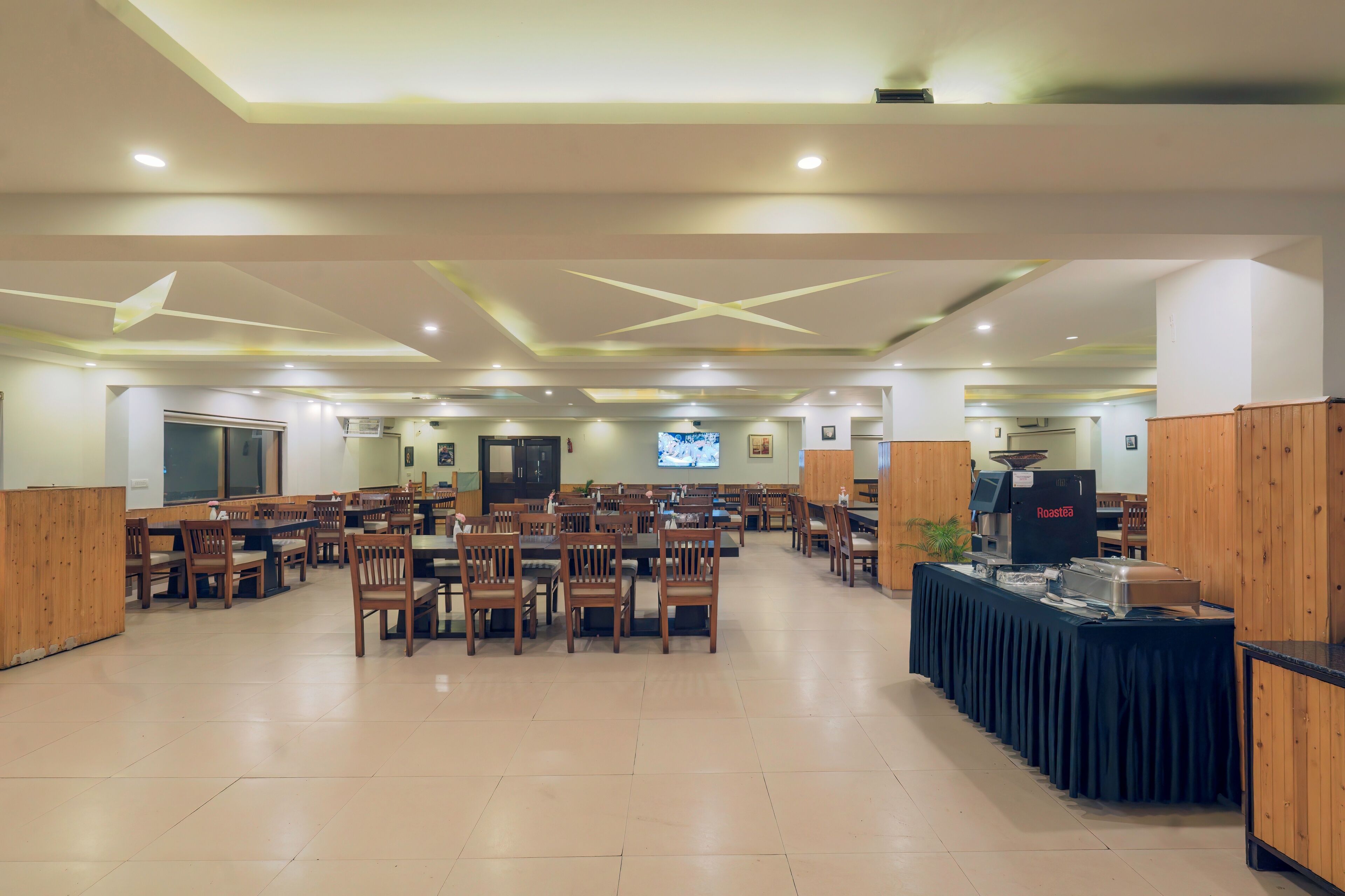A spacious dining area with multiple wooden tables and chairs, a buffet station, and a television screen in the background.