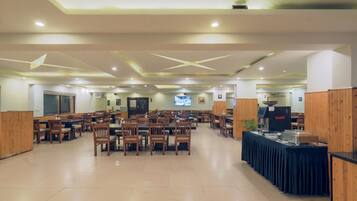 A spacious dining area with multiple wooden tables and chairs, a buffet station, and a television screen in the background.