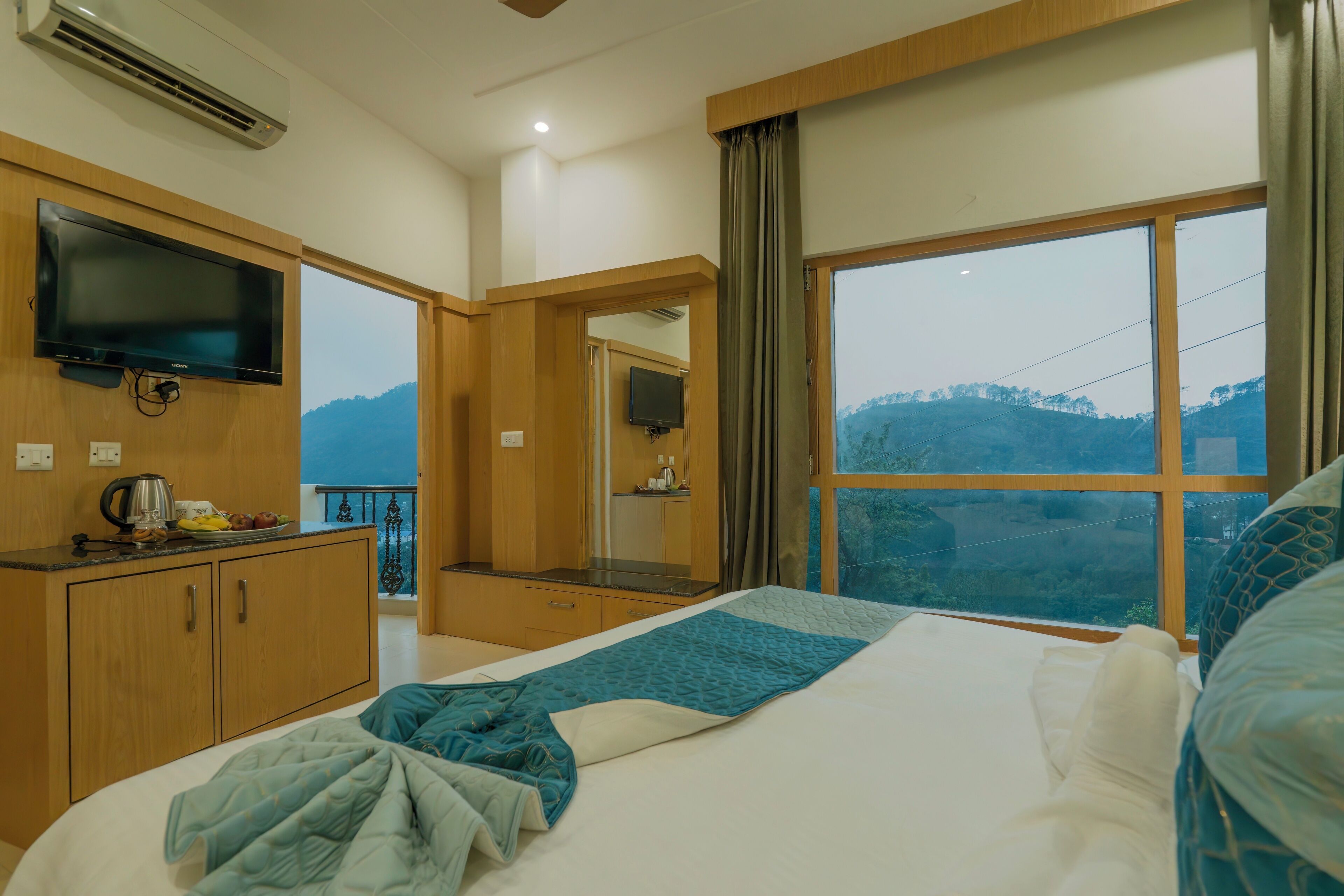 A hotel room with a bed, a TV, a window with a view of the mountains, and a kitchenette area.